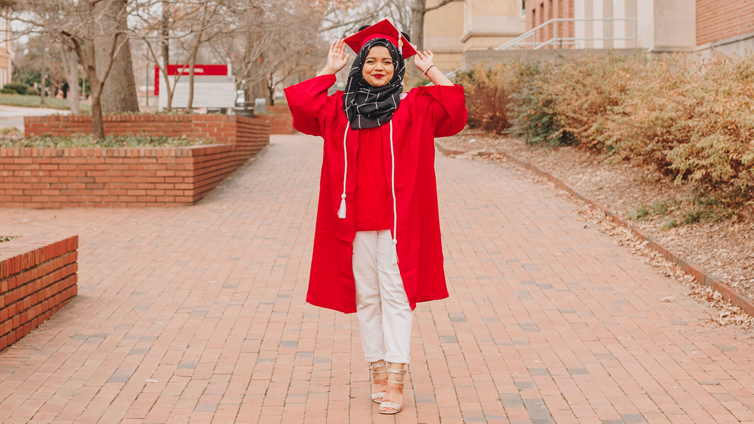 Wearing a red cap and gown, a student stands on a brick path