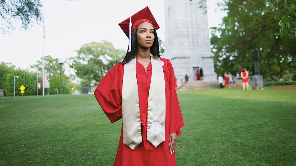 Ruhama Wolle stands in her red cap and gown in front of the NC State belltower
