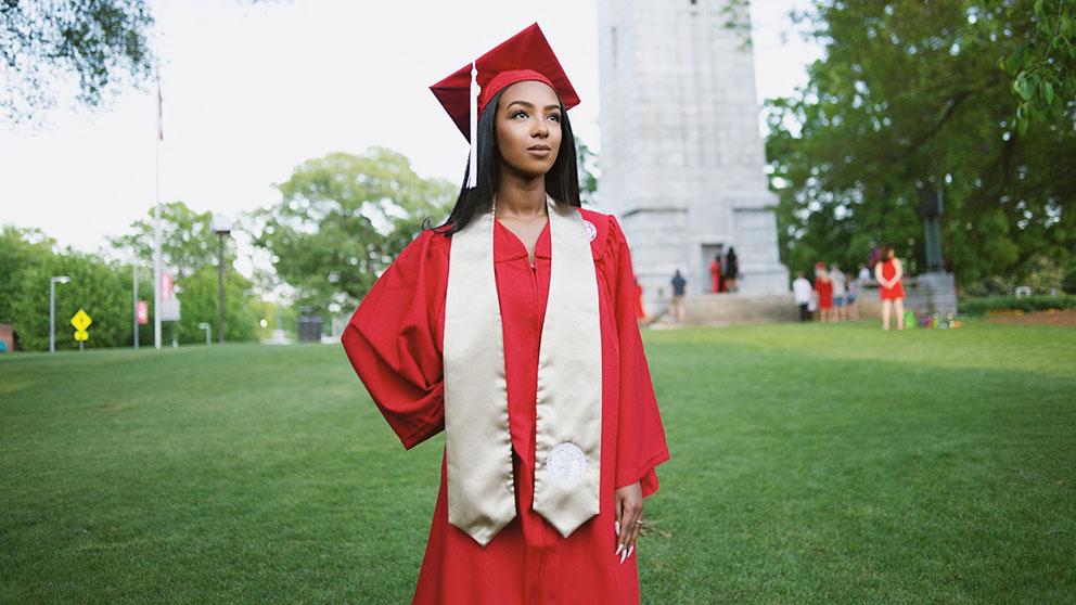 A student stands in her cap and gown in front of the NC State belltower.