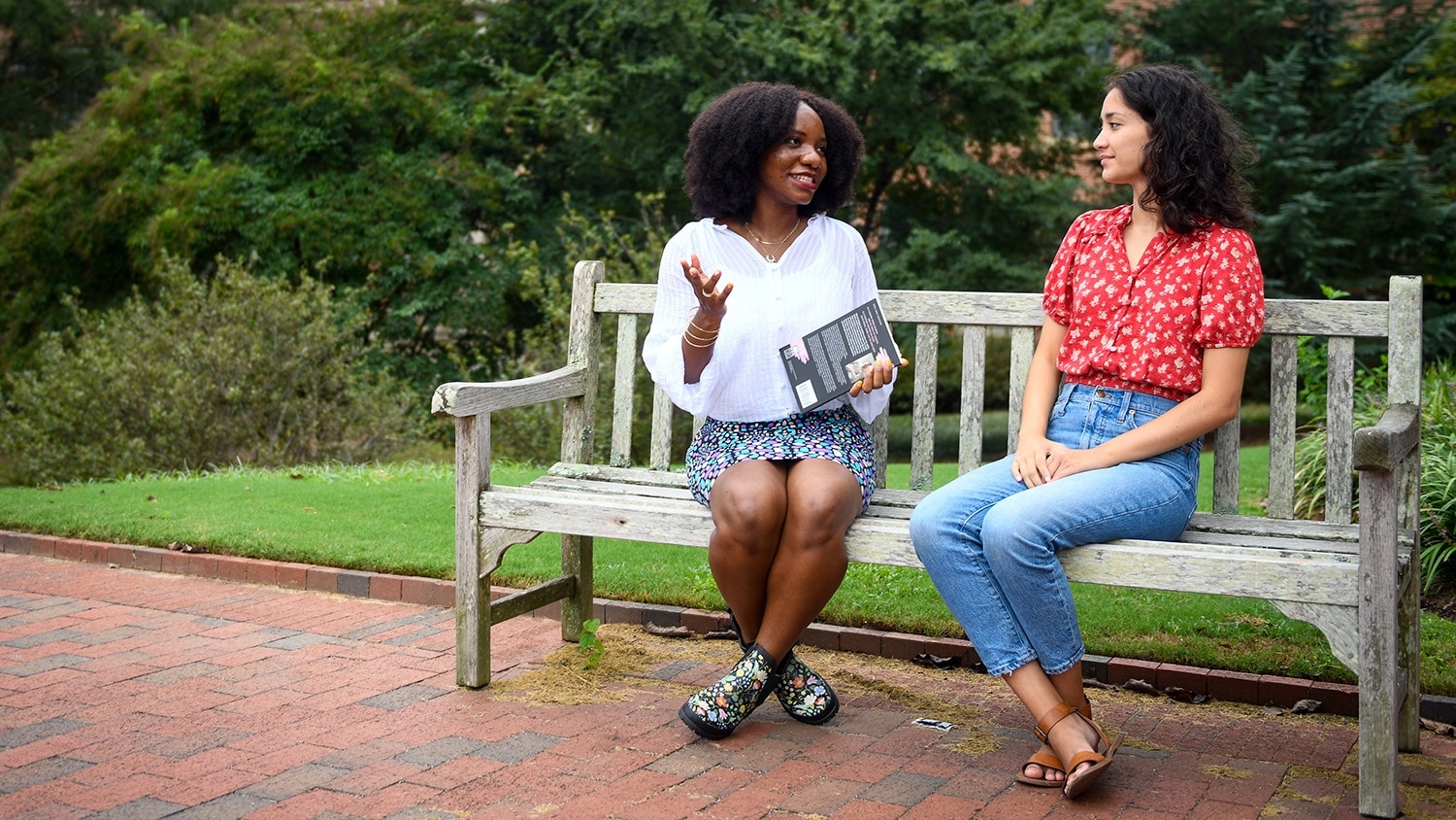 Rafeeat Aliyu and Meghan Tanaka sit beside each other on a bench on campus and discuss creative writing.