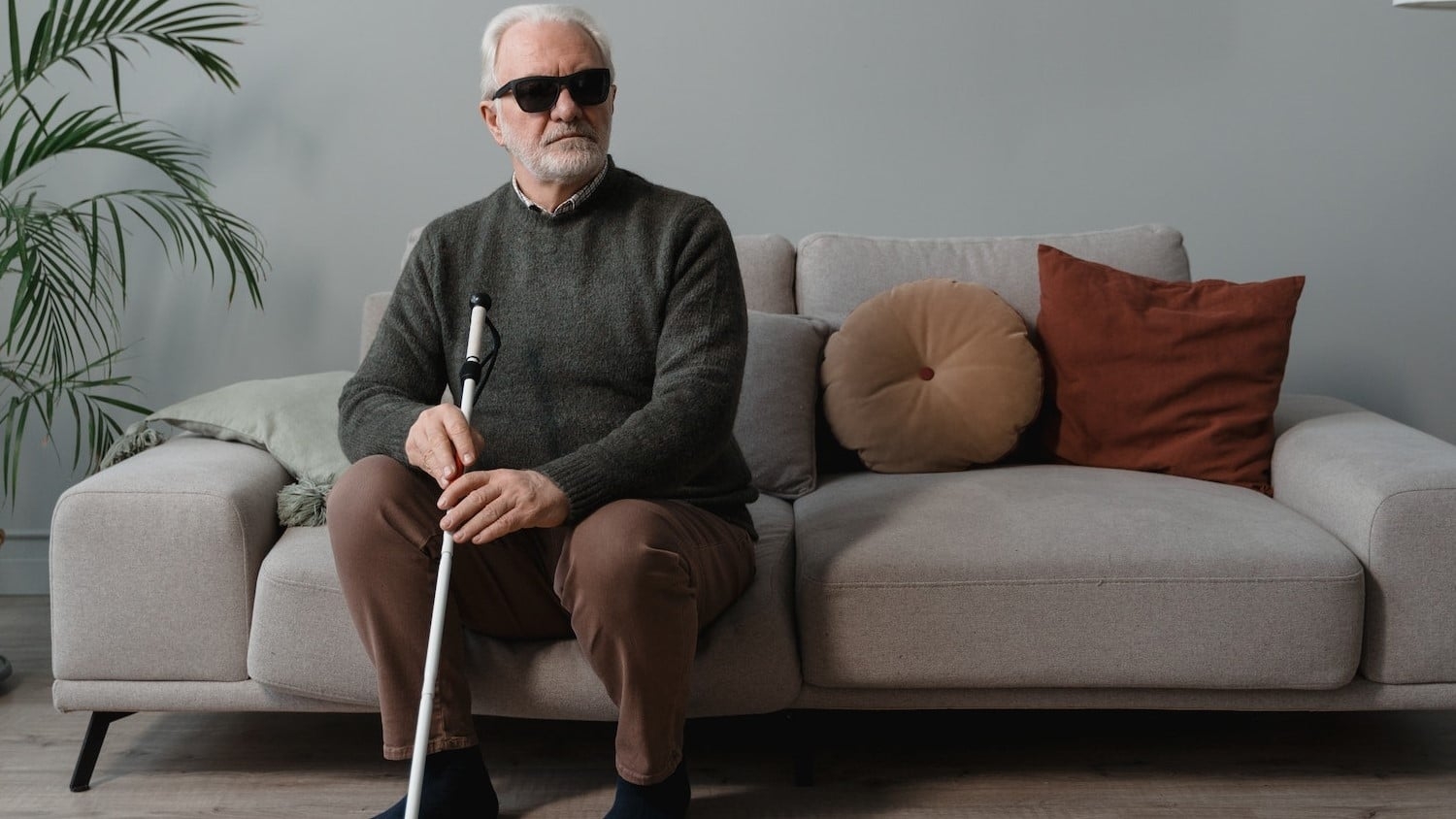 an older white man wearing sunglasses and holding a white cane is seated on a sofa