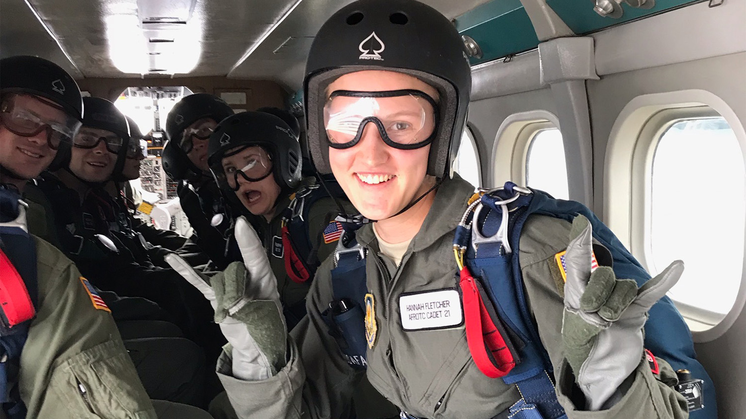Hannah Fletcher dressed in paratrooper gear in the back of a plane holds up her hands in wolfie signs