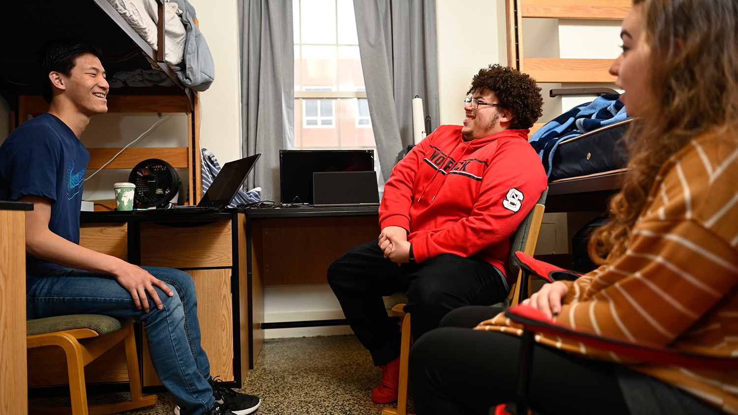 Three students talk and laugh while sitting in a dorm room.