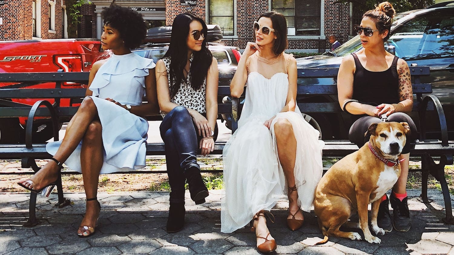 four young women of different races sit on a bench on a busy street.