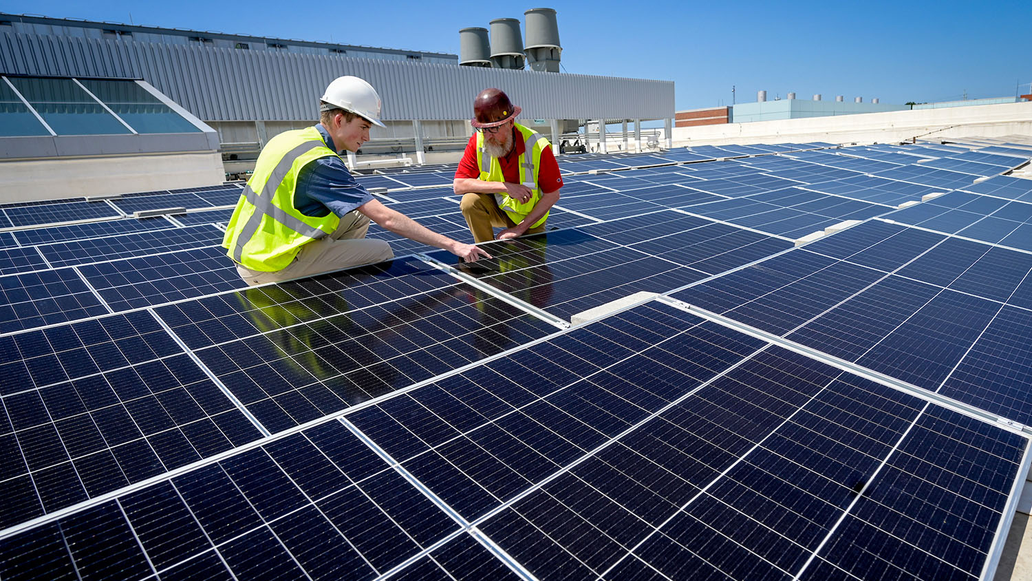 two men in hard hats crouch on a large roof covered with solar panels