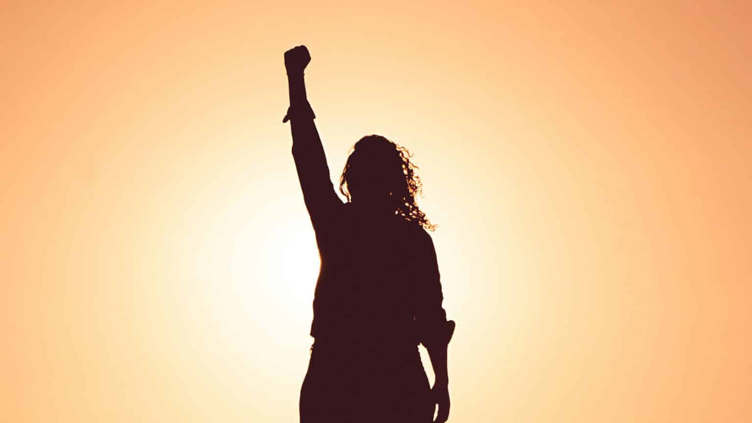 A shadowed woman holds her fist up over her head triumphantly.